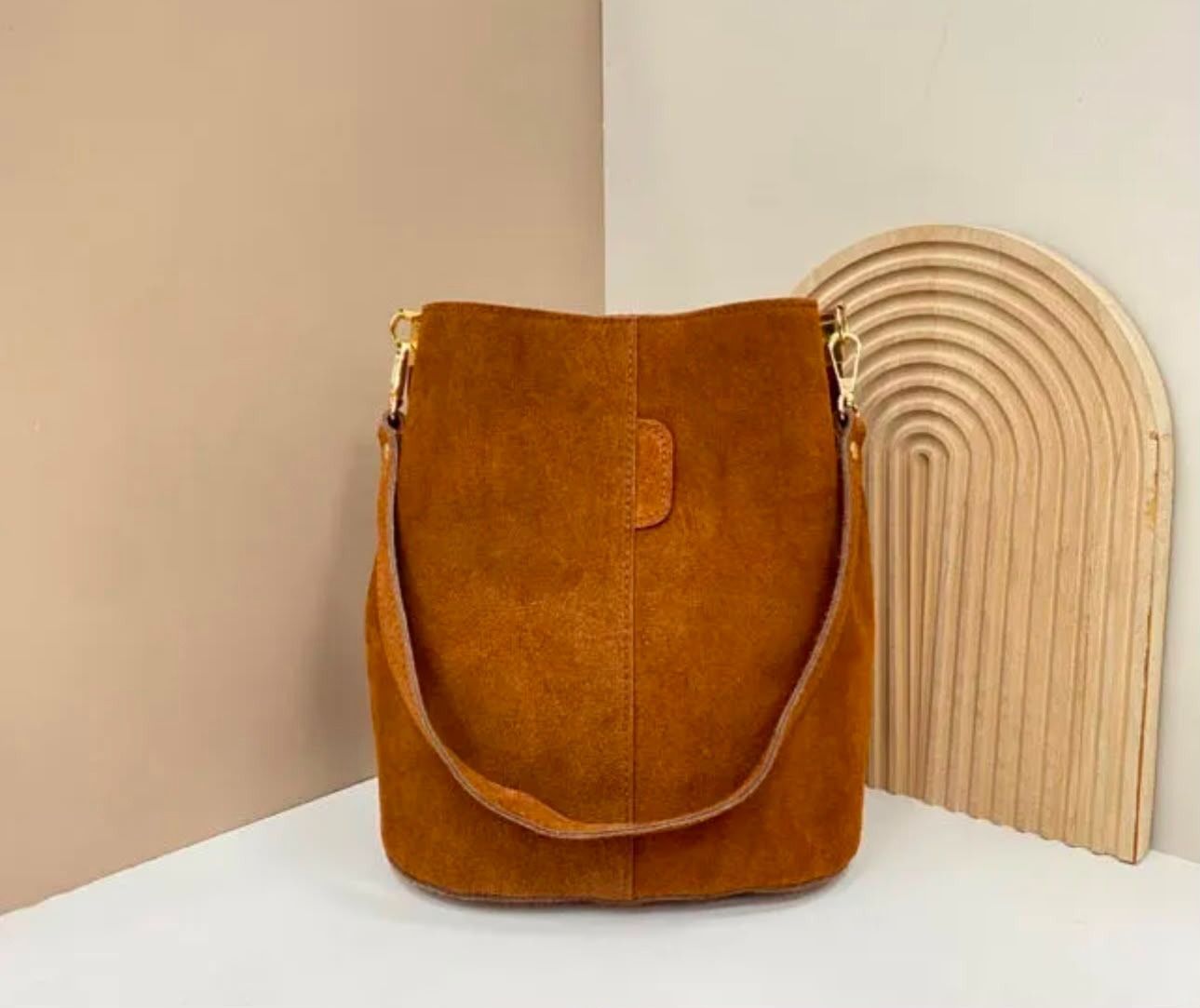Bolso bandolera de ante marrón sobre una superficie blanca junto a un arco decorativo de madera.