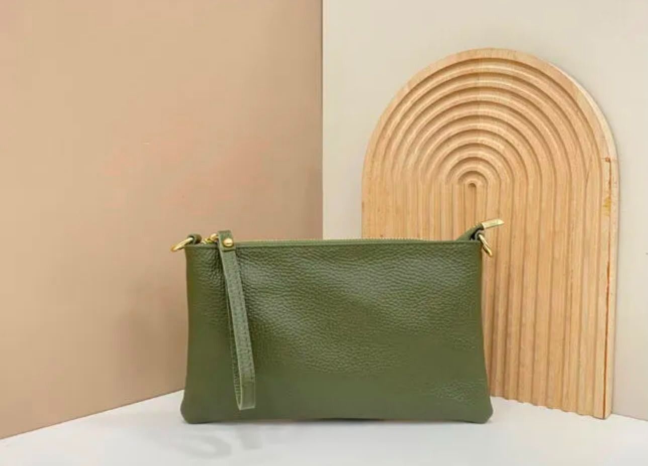 Un bolso de mano verde con cremallera sobre una superficie blanca, junto a un arco decorativo de madera.
