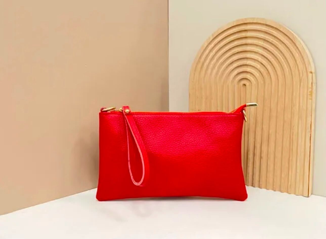 Un bolso de mano rojo con correa para la muñeca sobre una superficie blanca junto a un arco decorativo de madera.