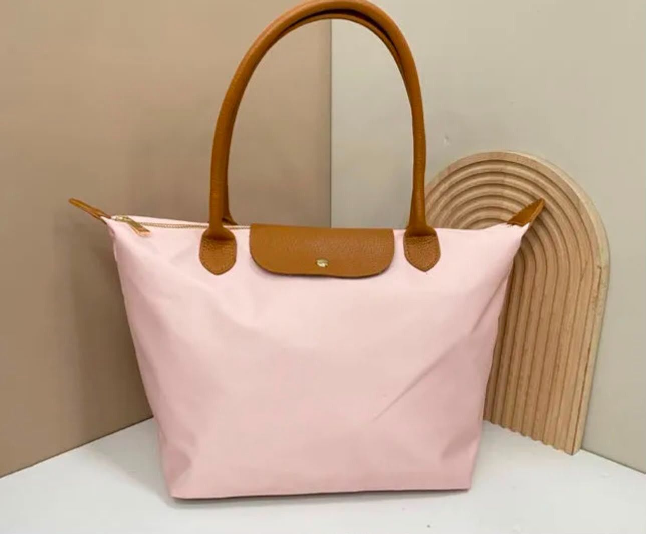 Bolso tote rosa claro con asas y ribetes color tostado sobre una superficie blanca, contra una pared beige.