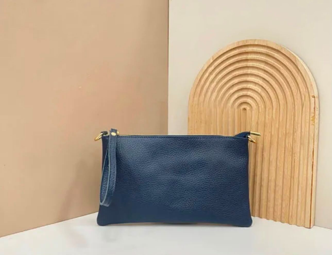 Un bolso de cuero azul sobre una superficie blanca junto a una pieza decorativa de madera arqueada contra una pared beige.