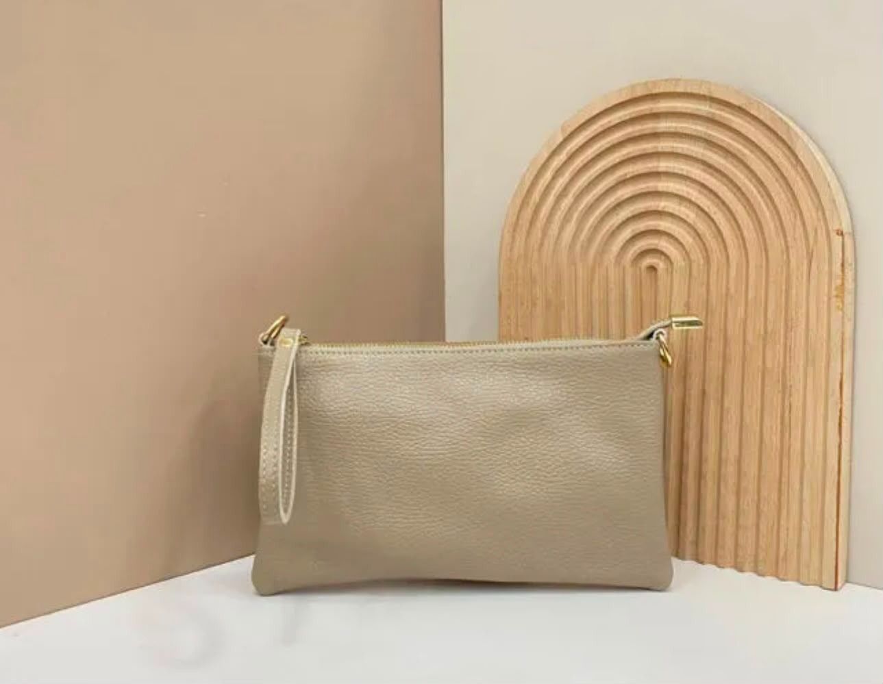 Un bolso de mano beige con correa para la muñeca sobre una superficie blanca junto a una pieza decorativa de madera curvada.