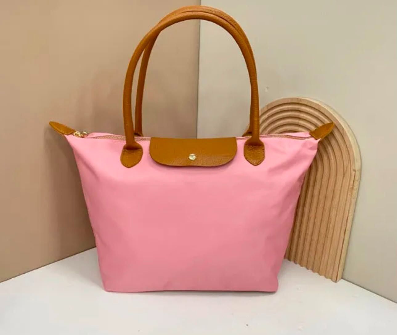 Bolso tote rosa con asas y solapa color camel, mostrado sobre una pared blanca con un fondo tejido en forma de abanico.
