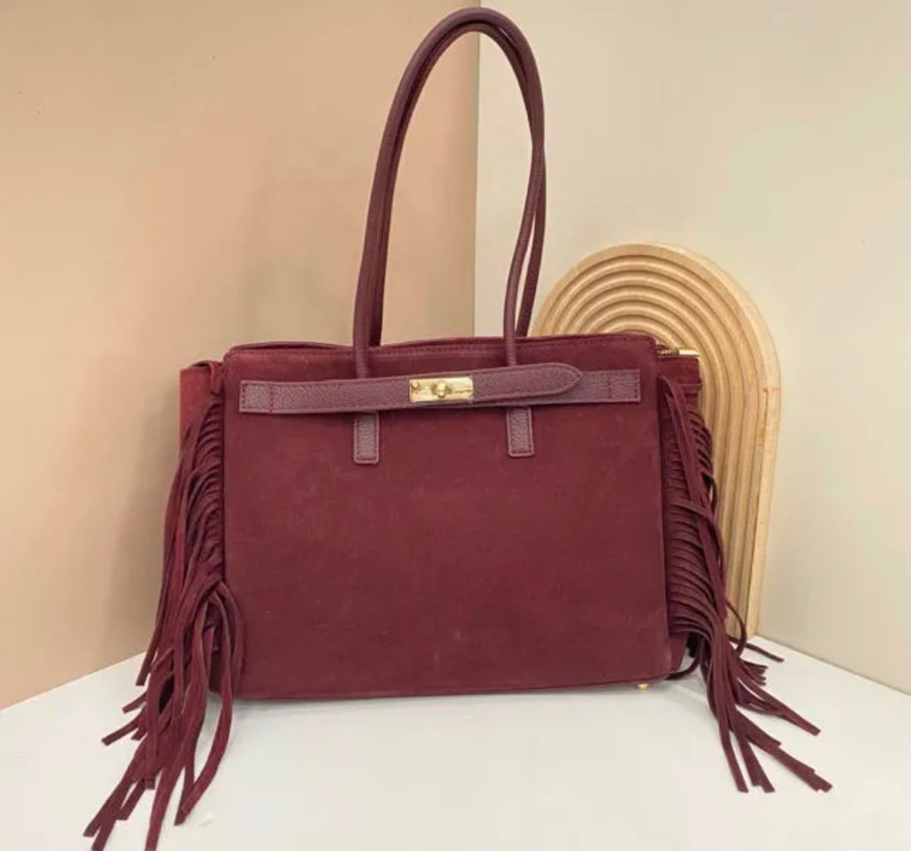 Bolso de mano de ante color burdeos con flecos, mostrado sobre una superficie blanca con fondo beige.