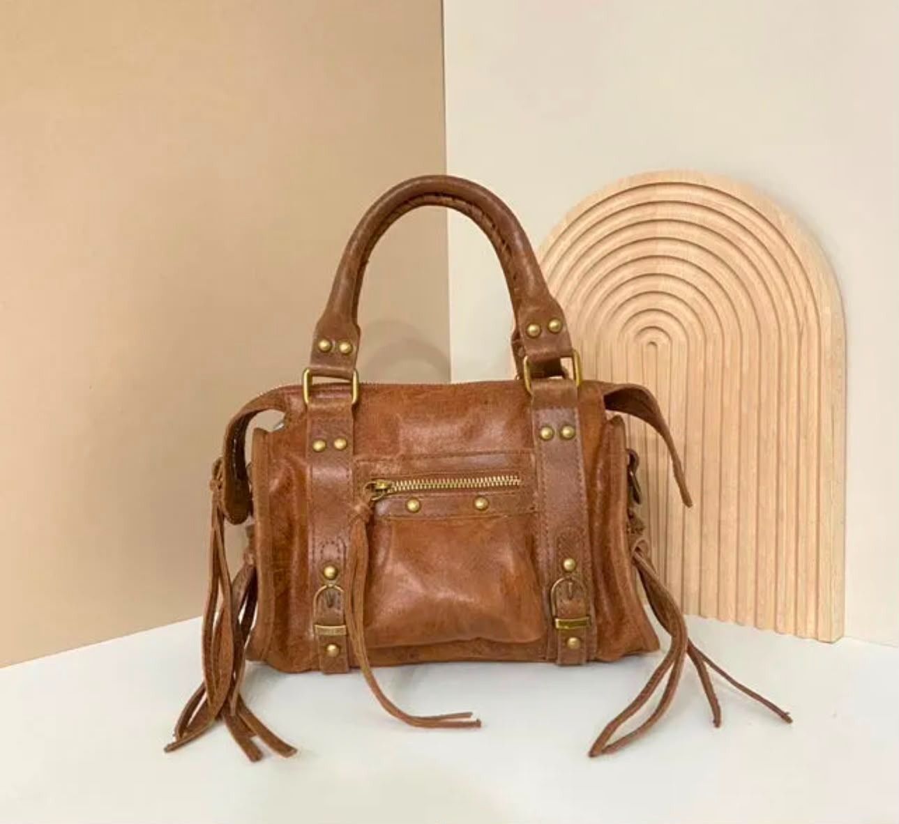 Bolso de piel marrón con borlas en un estante junto a un arco decorativo de madera clara.