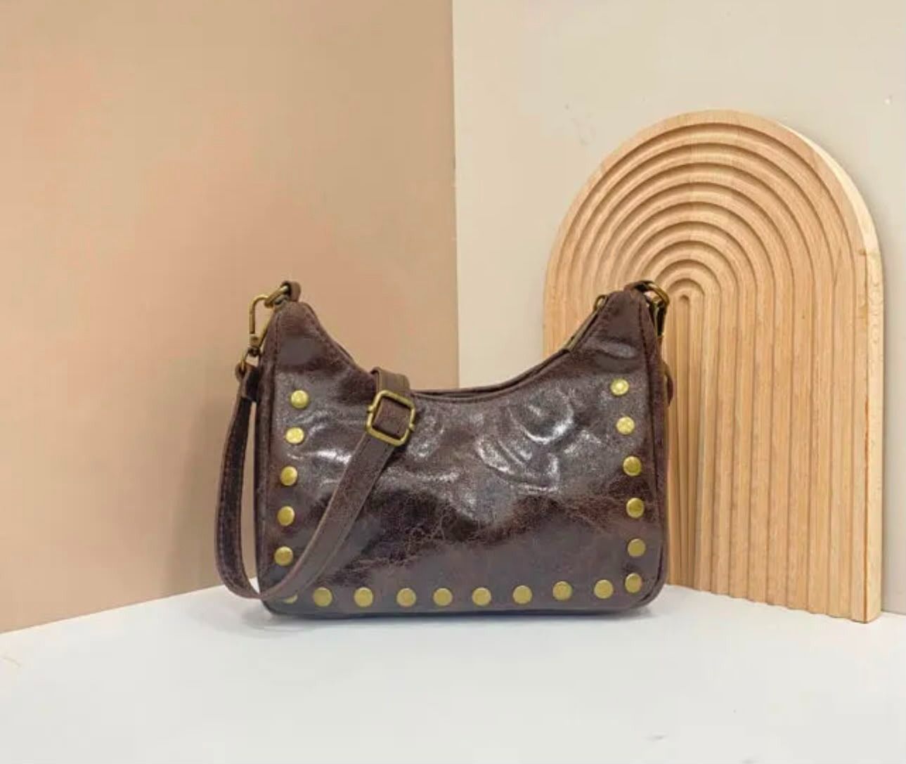 Bolso de hombro de piel marrón con tachuelas doradas junto a un arco decorativo de madera sobre una superficie blanca.