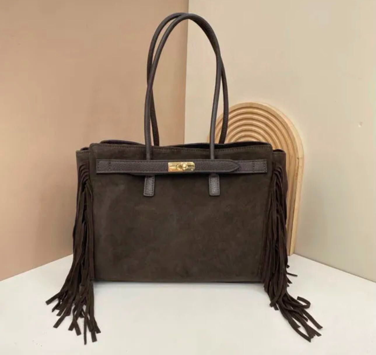 Bolso de mano de ante marrón con flecos y asas negras sobre una superficie blanca.
