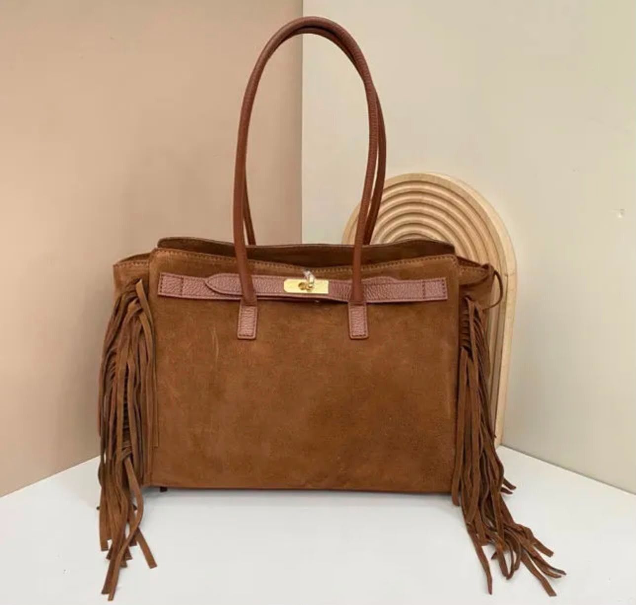 Bolso de mano de ante marrón con asas largas y flecos, sobre un fondo beige.