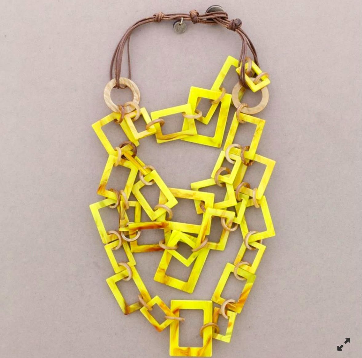 Collar geométrico amarillo con eslabones cuadrados sobre un cordón marrón sobre un fondo beige.