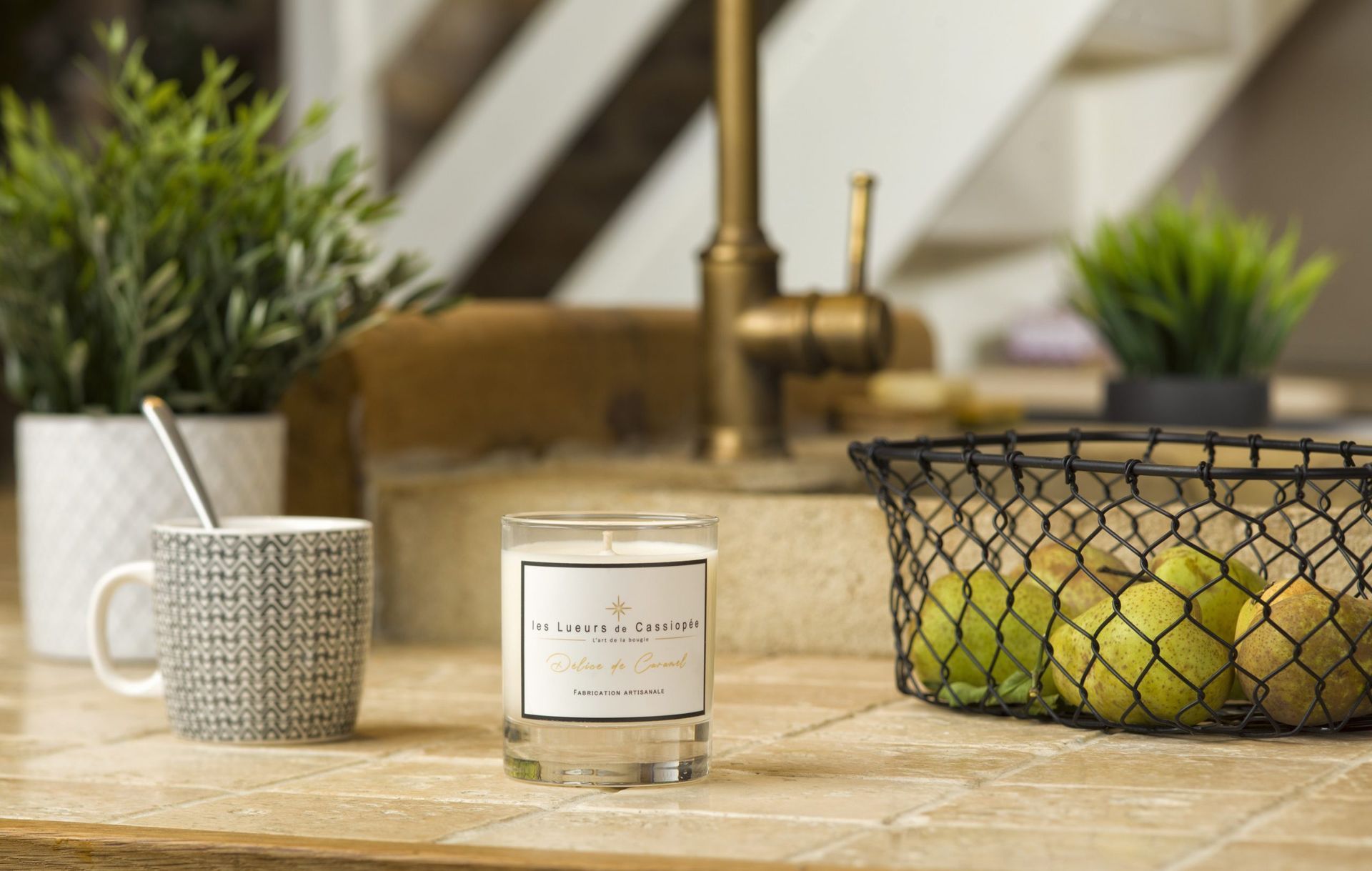 Un pot à bougie, une tasse en métal, des herbes aromatiques en pot et une corbeille de fruits sont posés sur un plan de travail en pierre près de l'évier de la cuisine.