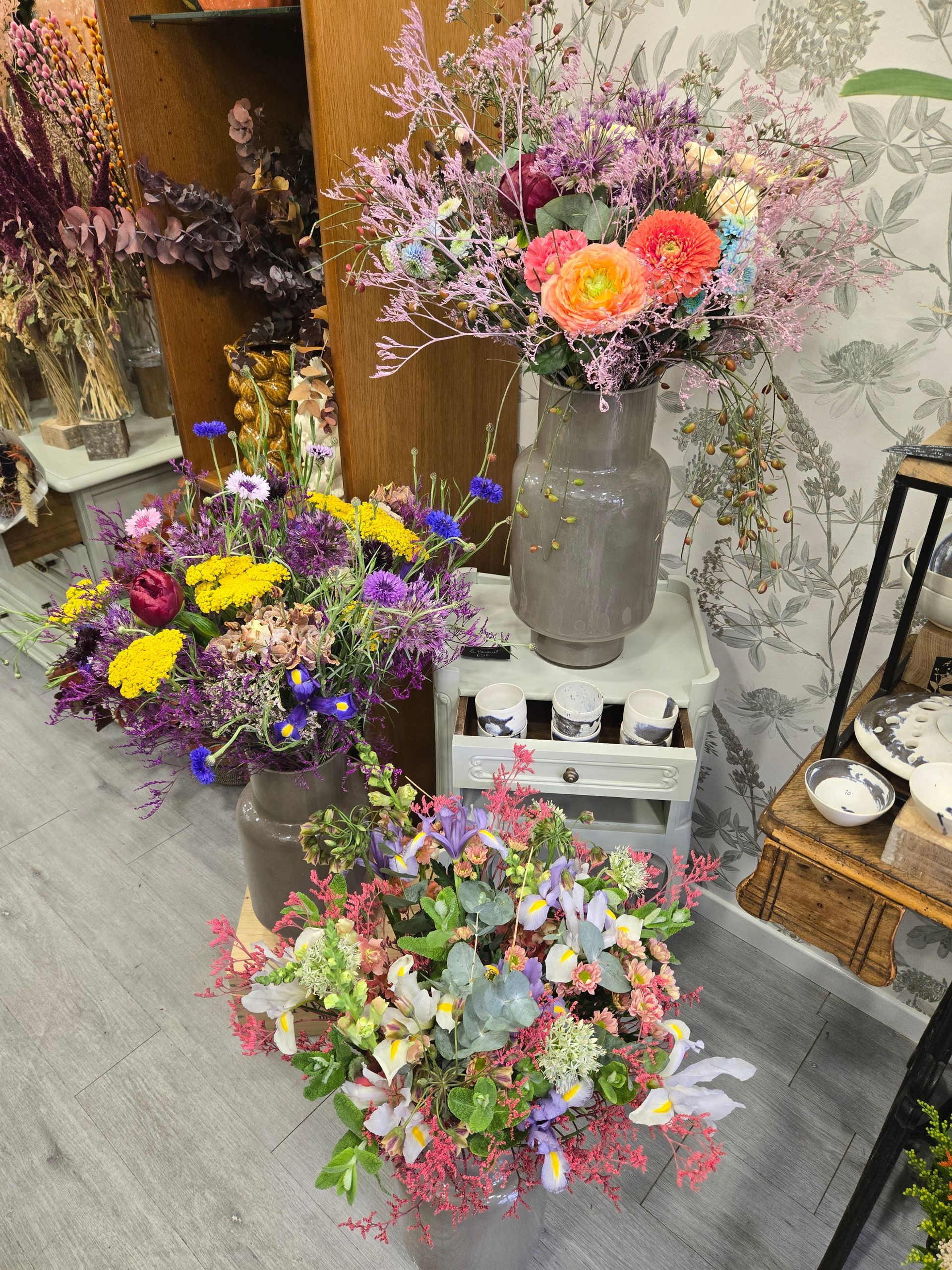 Présentation de fleurs avec des bouquets colorés dans un magasin, dont un bouquet rose dans un grand vase gris posé sur des caisses blanches.
