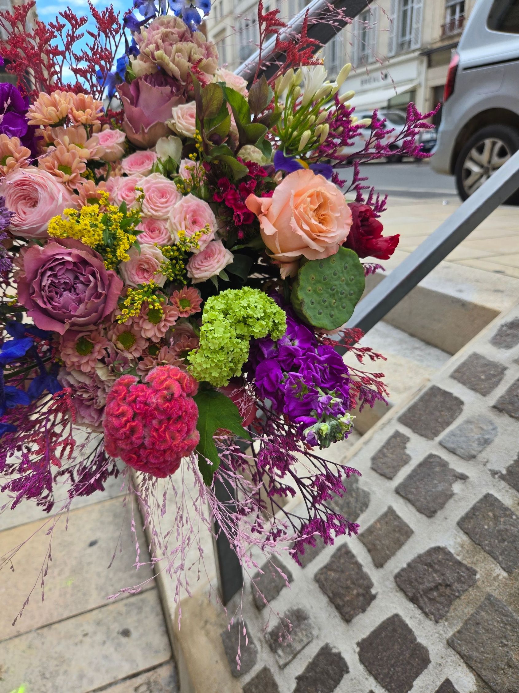 Bouquet coloré de fleurs roses, violettes, pêche et vertes, posé sur un trottoir en bordure de rue.