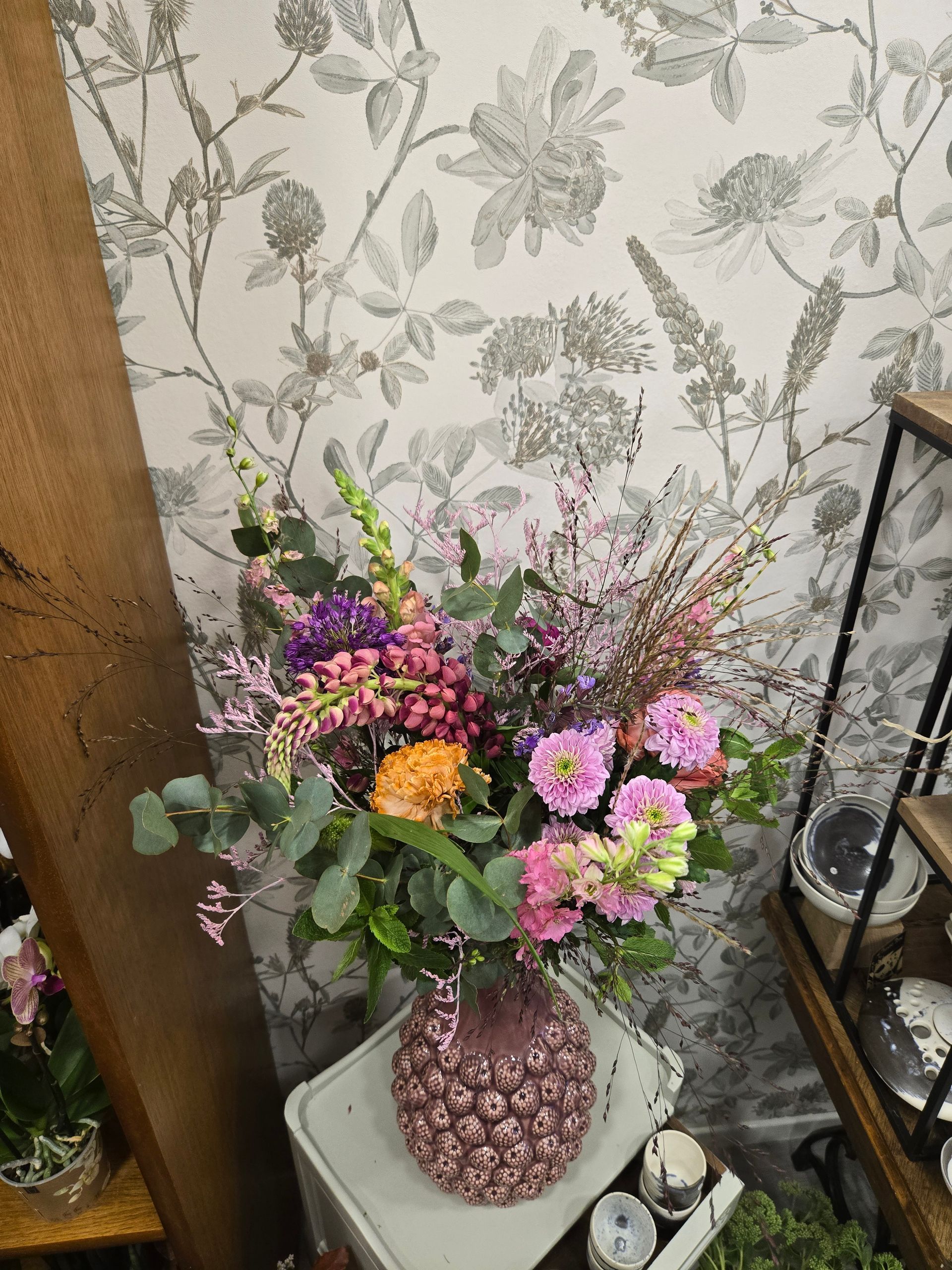 Bouquet coloré de fleurs variées dans un vase à motifs sur une table à côté d'un mur tapissé de papier peint à fleurs