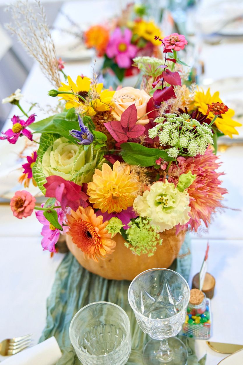 Un bouquet de fleurs variées et colorées dans un vase, posé sur une table avec de la verrerie et une petite bouteille.