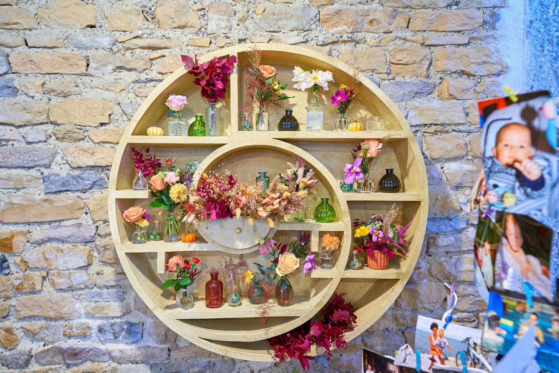 Étagère murale circulaire en bois remplie de fleurs colorées et de petits objets décoratifs, adossée à un mur en pierre.
