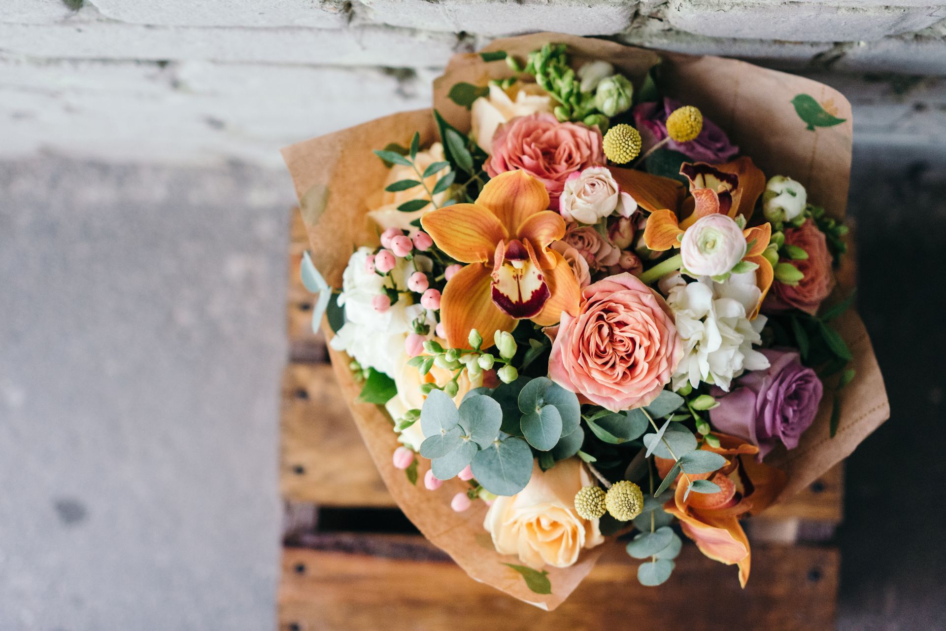 Bouquet de fleurs mélangées dans les tons pêche, rose, blanc et violet, emballé dans du papier kraft et placé sur une caisse en bois