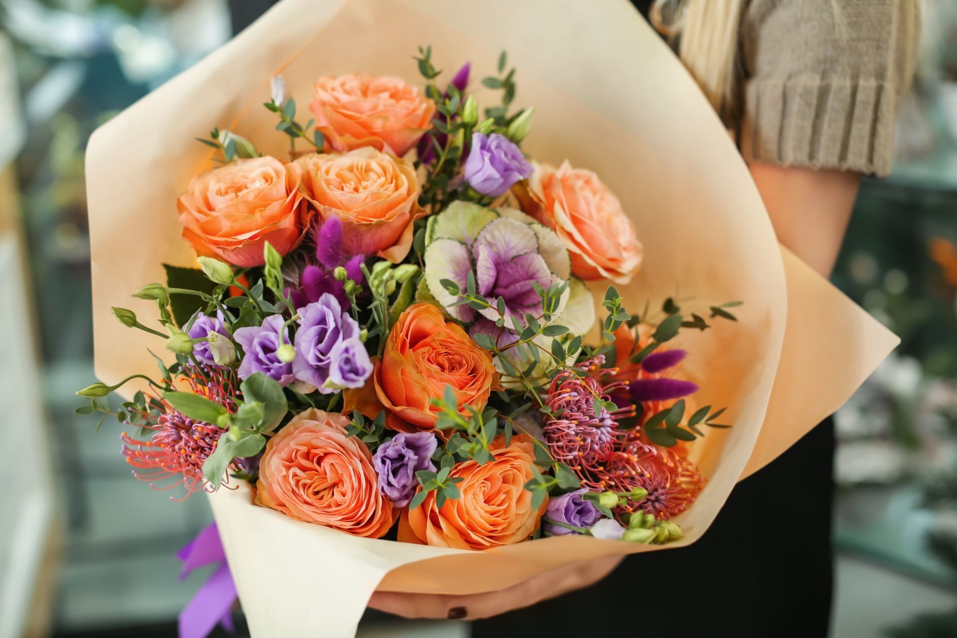 Bouquet de roses orange et de fleurs violettes enveloppé dans du papier beige, tenu par une personne