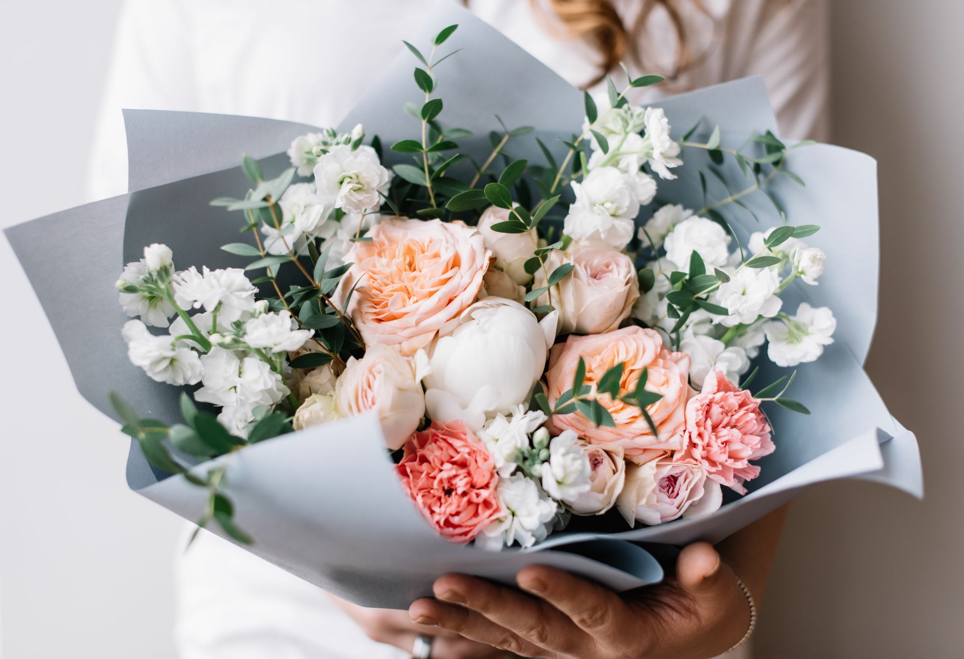 Bouquet de fleurs rose pastel et blanches enveloppé dans du papier gris, tenu dans les mains