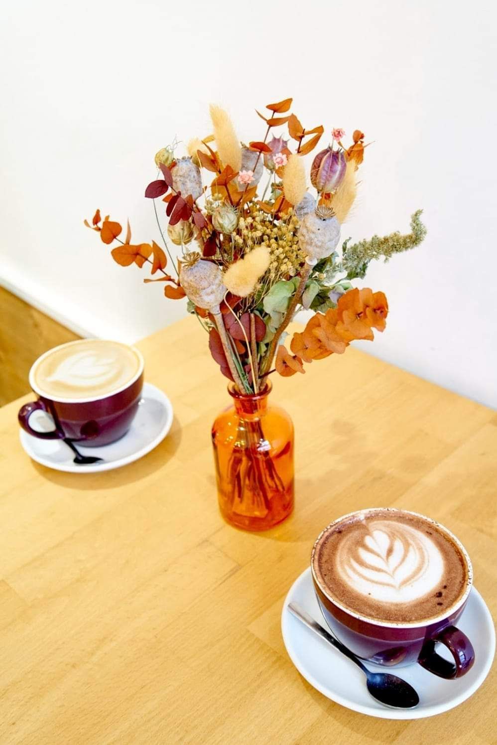 Deux tasses à café sur leurs soucoupes, à côté d'un vase orange de fleurs séchées, sur une table en bois.