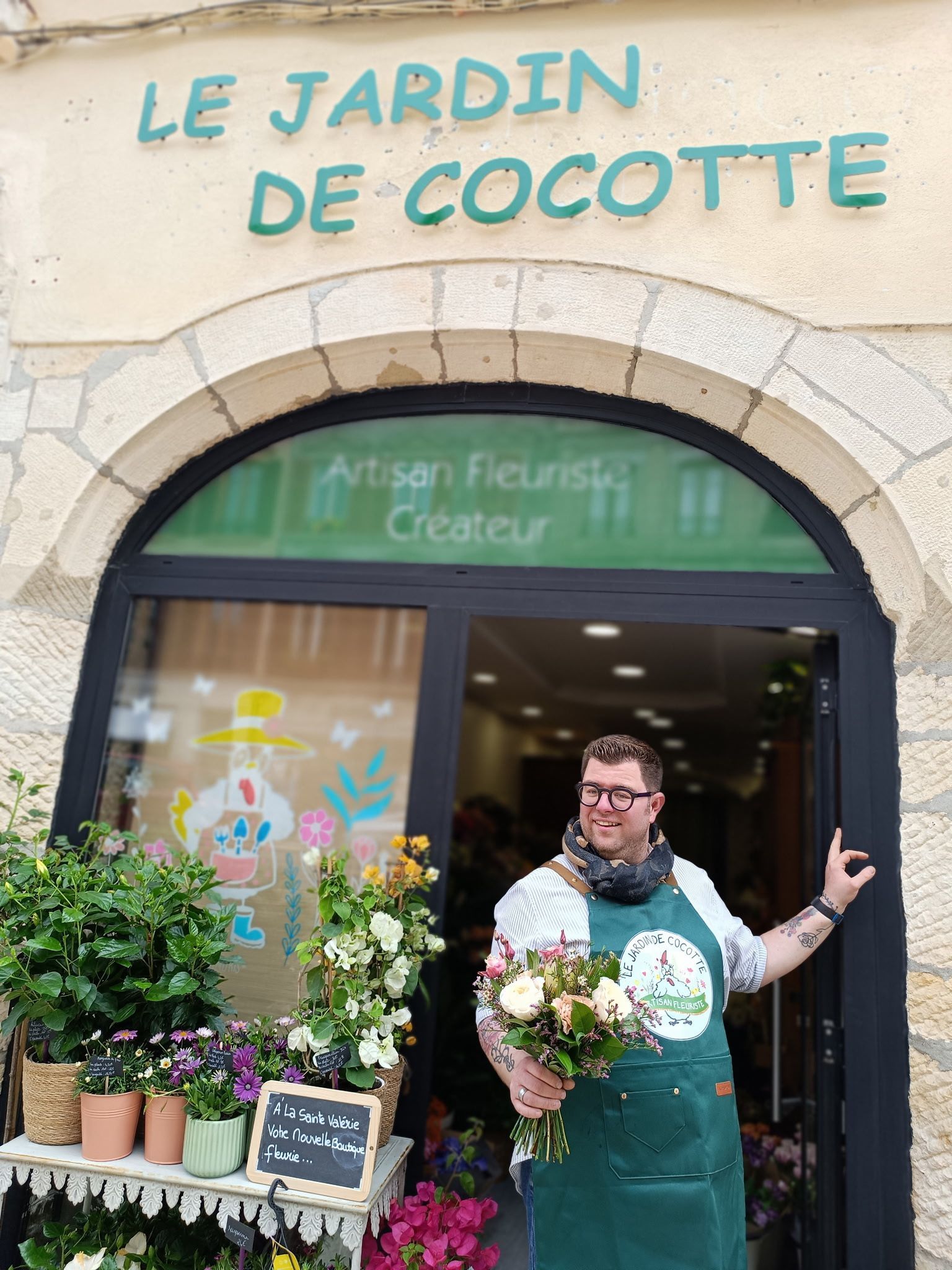 Une personne en tablier vert tenant des fleurs devant l'enseigne du magasin.
