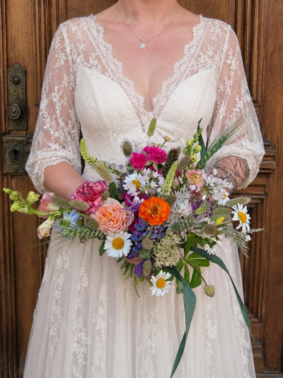 Une mariée en robe de mariée en dentelle tenant un bouquet de fleurs sauvages colorées devant des portes en bois