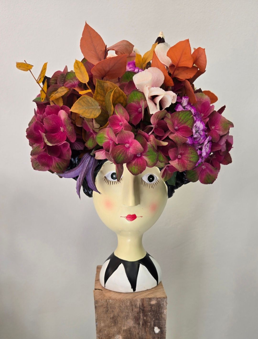 Tête de poupée recouverte de fleurs colorées et visage sculpté sur un socle en bois