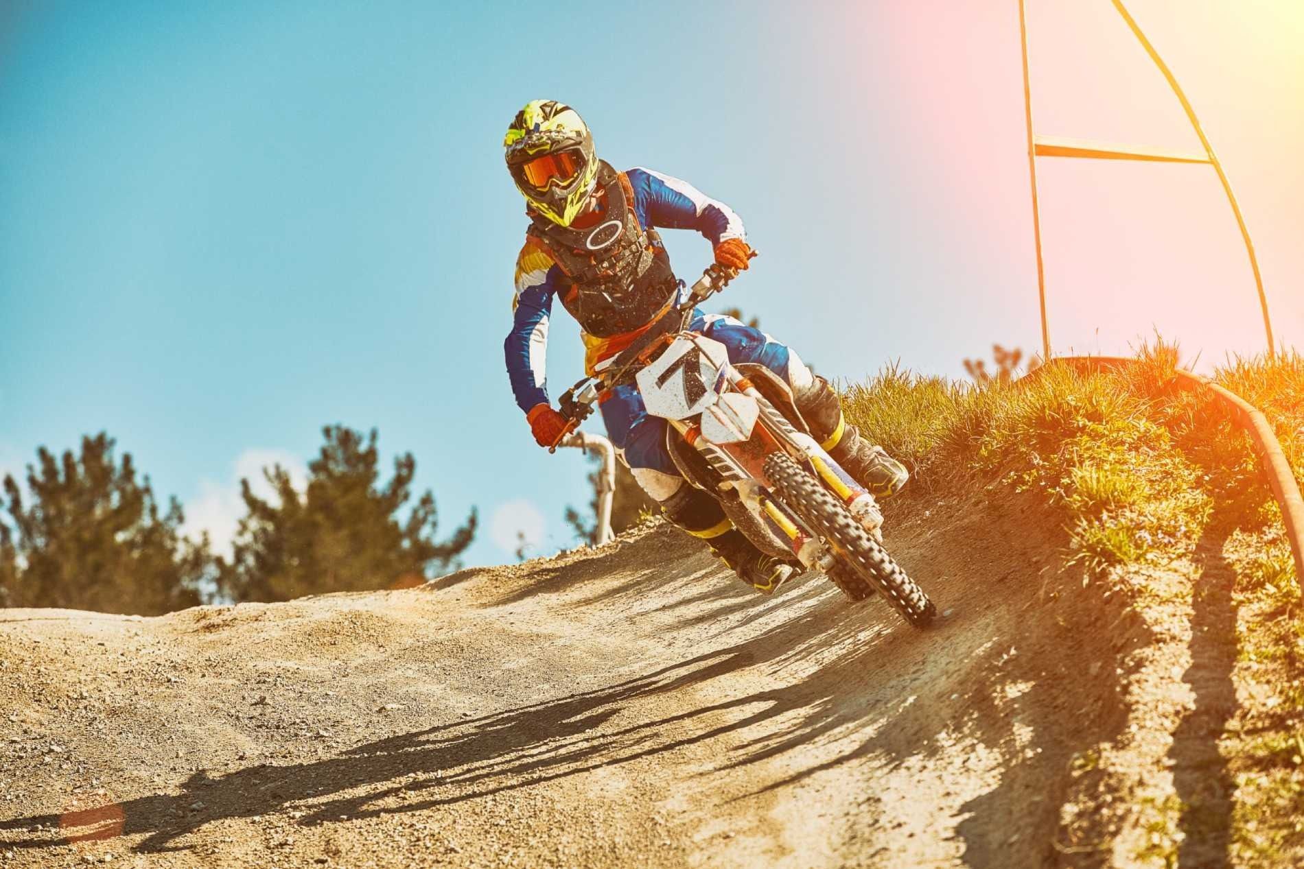 Un piloto de motocross con casco blanco y equipación roja está sentado en una moto de cross, levantando una mano enguantada cerca de una furgoneta.