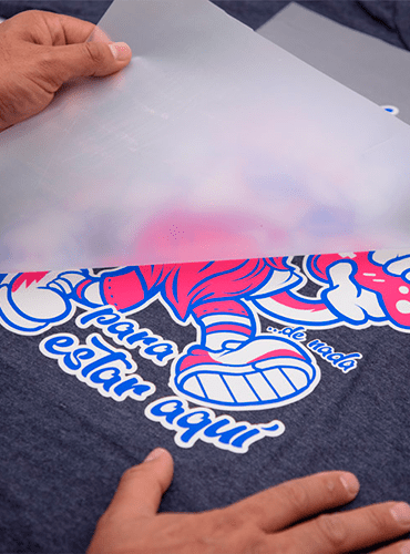 Persona pelando papel de transferencia de un gráfico serigrafiado en una camiseta gris oscuro.