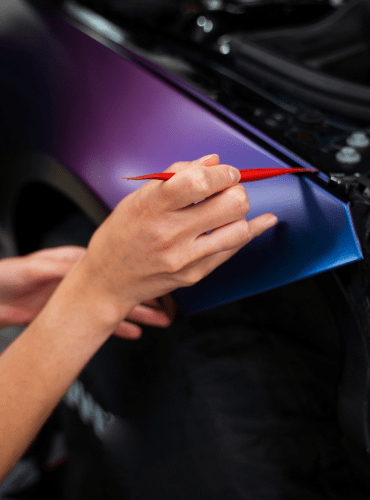 Persona que utiliza una herramienta para aplicar un revestimiento de color azul/violeta al guardabarros de un vehículo negro.