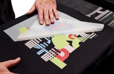 Persona pelando una hoja transparente de una camiseta negra con un diseño abstracto y colorido.