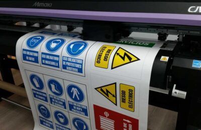 Una impresora de gran formato que imprime señales de seguridad con símbolos azules, amarillos y rojos en papel blanco.
