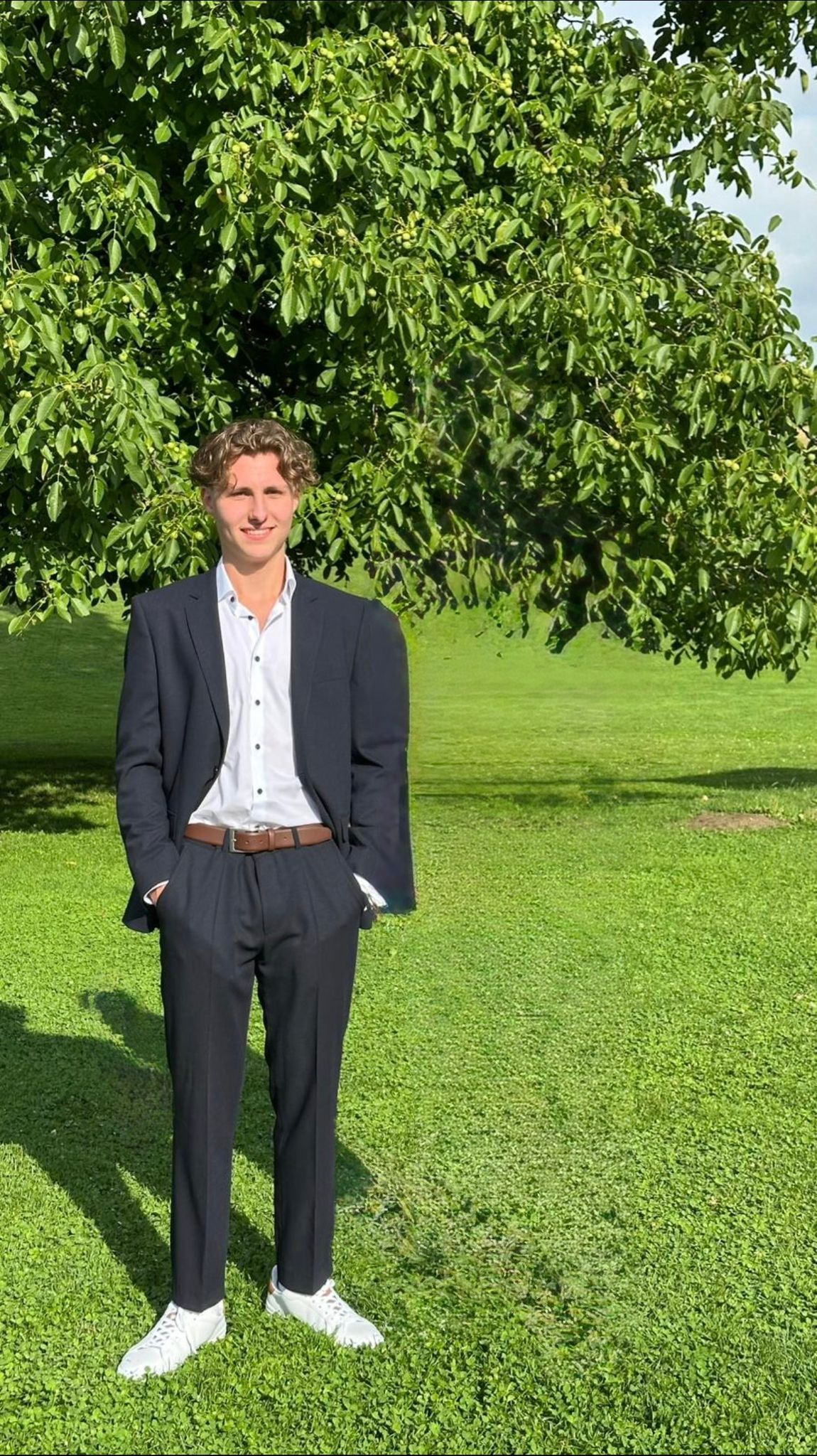 Mann in dunklem Anzug, weißem Hemd und Turnschuhen, steht mit den Händen in den Taschen auf einer Wiese vor einem Baum.