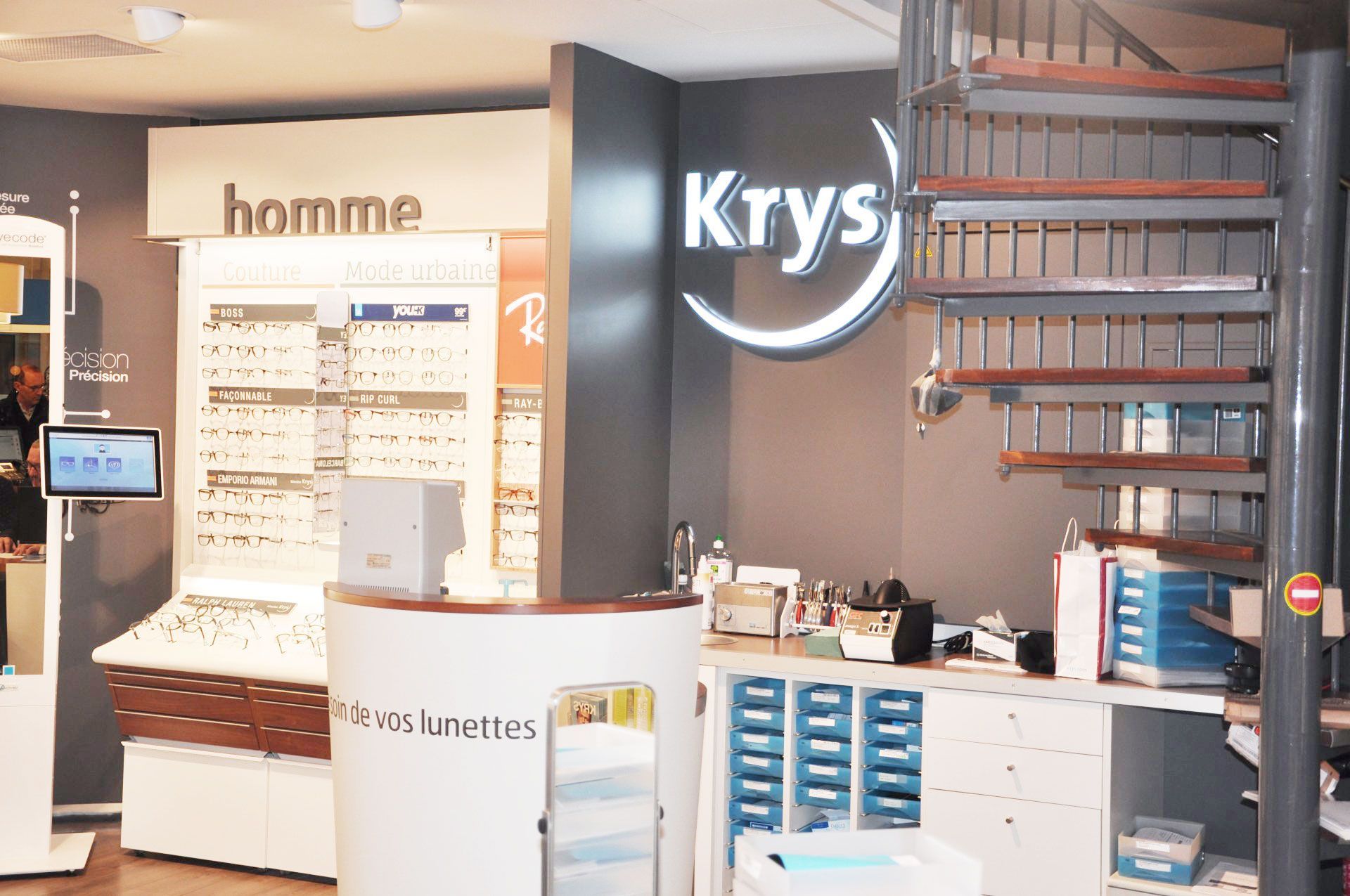 Intérieur du magasin d'Optique Pellé Krys avec des présentoirs de lunettes, un escalier en colimaçon et un comptoir blanc.