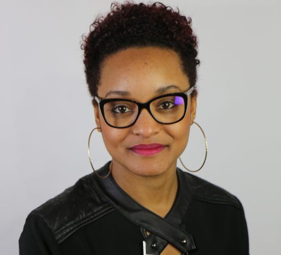 Femme aux cheveux courts et bouclés, portant des lunettes, des créoles et une veste noire, souriante.