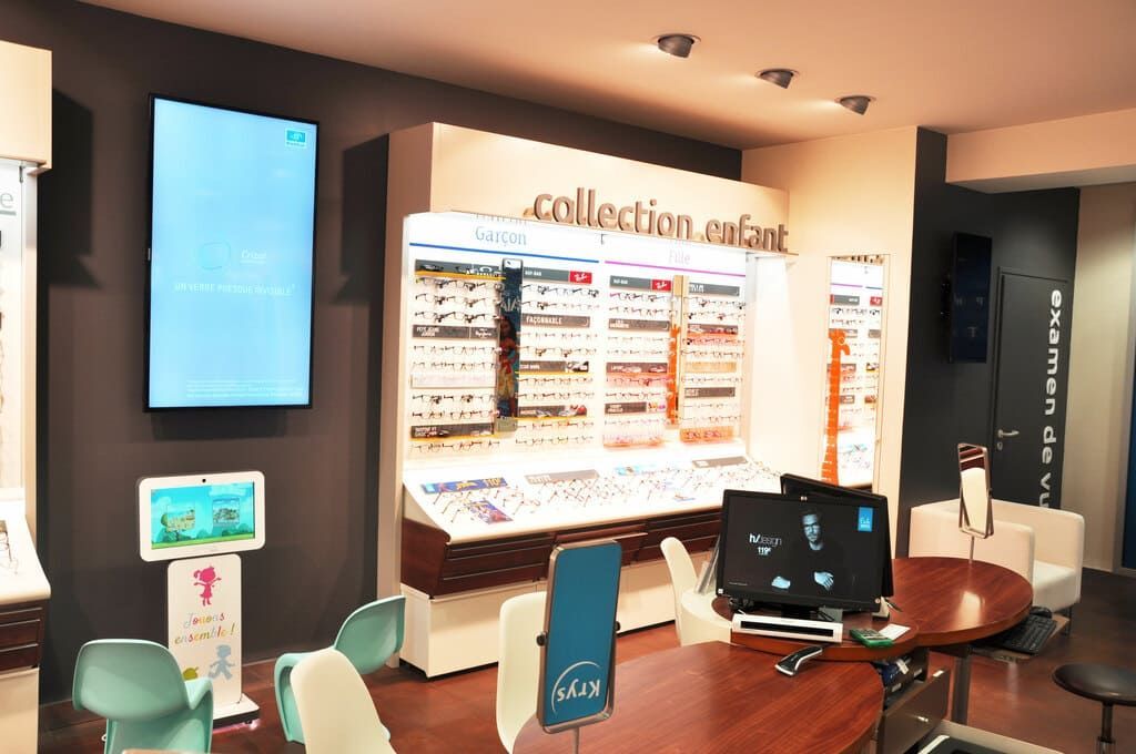Intérieur du magasin de lunettes Optique Pellé Krys, présentant des montures et des écrans numériques, avec des espaces pour s'asseoir.