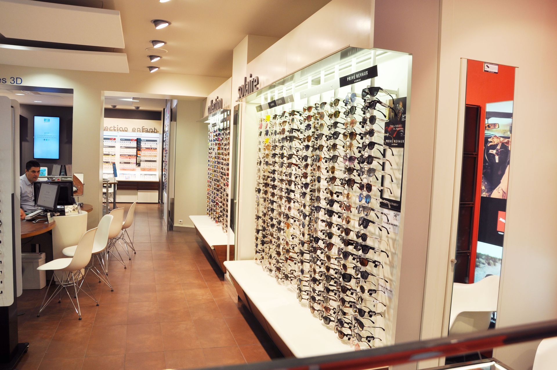 Intérieur d'un magasin d'optique avec des lunettes exposées aux murs. Une personne est assise à un bureau.