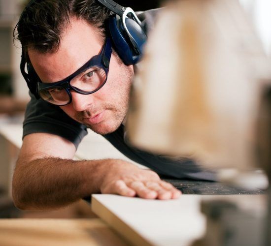 Un homme utilise une scie pour couper du bois, portant des lunettes de sécurité et une protection auditive.