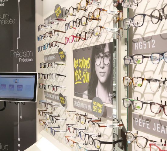 Des lunettes sont exposées dans un magasin d'optométrie, avec différentes montures, un écran et une affiche publicitaire.