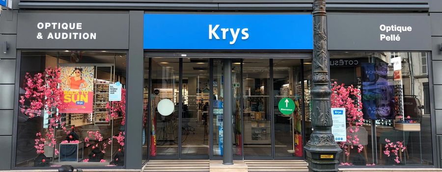 Une devanture portant l'enseigne Krys chez Optique Pellé. L'étalage présente des lunettes et des fleurs roses.