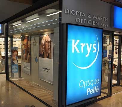 Extérieur du magasin d'Optique Pellé Krys, avec une enseigne bleue lumineuse et des vitrines présentant des lunettes et des publicités.