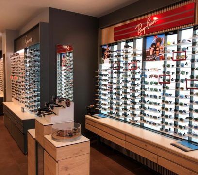 Intérieur d'un magasin d'optique présentant de nombreuses lunettes de soleil sur des étagères. Les tons boisés et gris dominent.