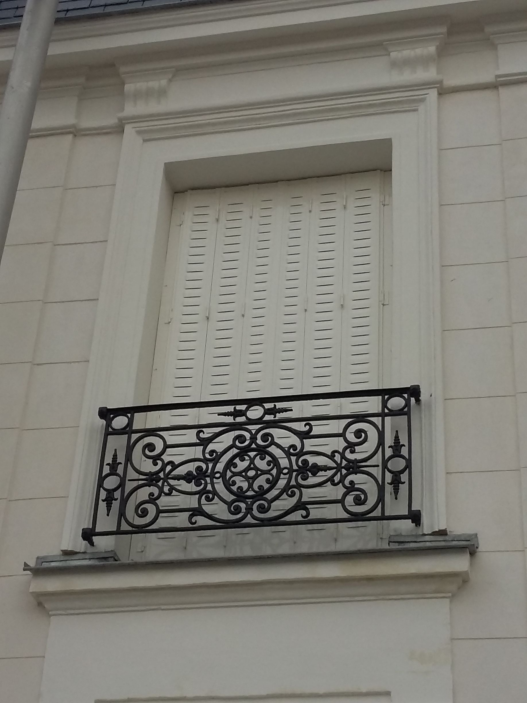 Fenêtre avec balcon décoratif en fer forgé. Bâtiment beige, volets blancs, détails ornementaux.