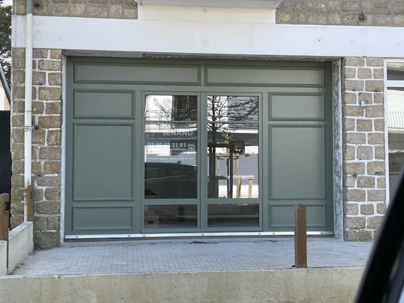 Façade de magasin à panneaux verts avec fenêtres en verre, située dans un bâtiment en pierre ; reflet de la vue sur la rue.
