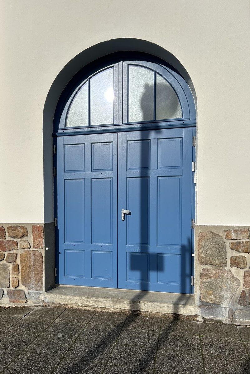 Porte double bleue avec fenêtre cintrée, encastrée dans un mur blanc avec base en pierre.