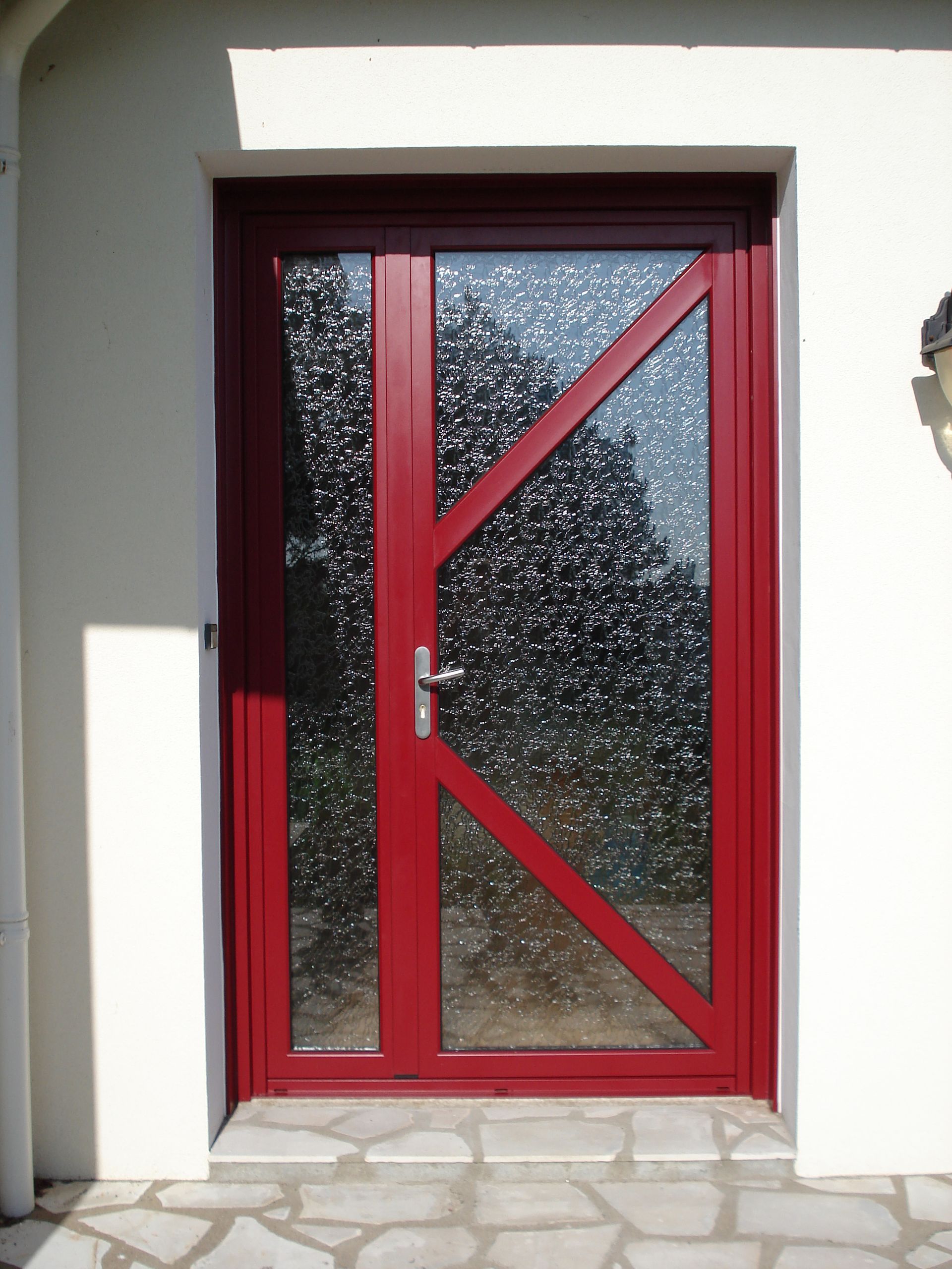 Porte en verre rouge avec supports inclinés, insérée dans un extérieur blanc avec des marches en pierre.