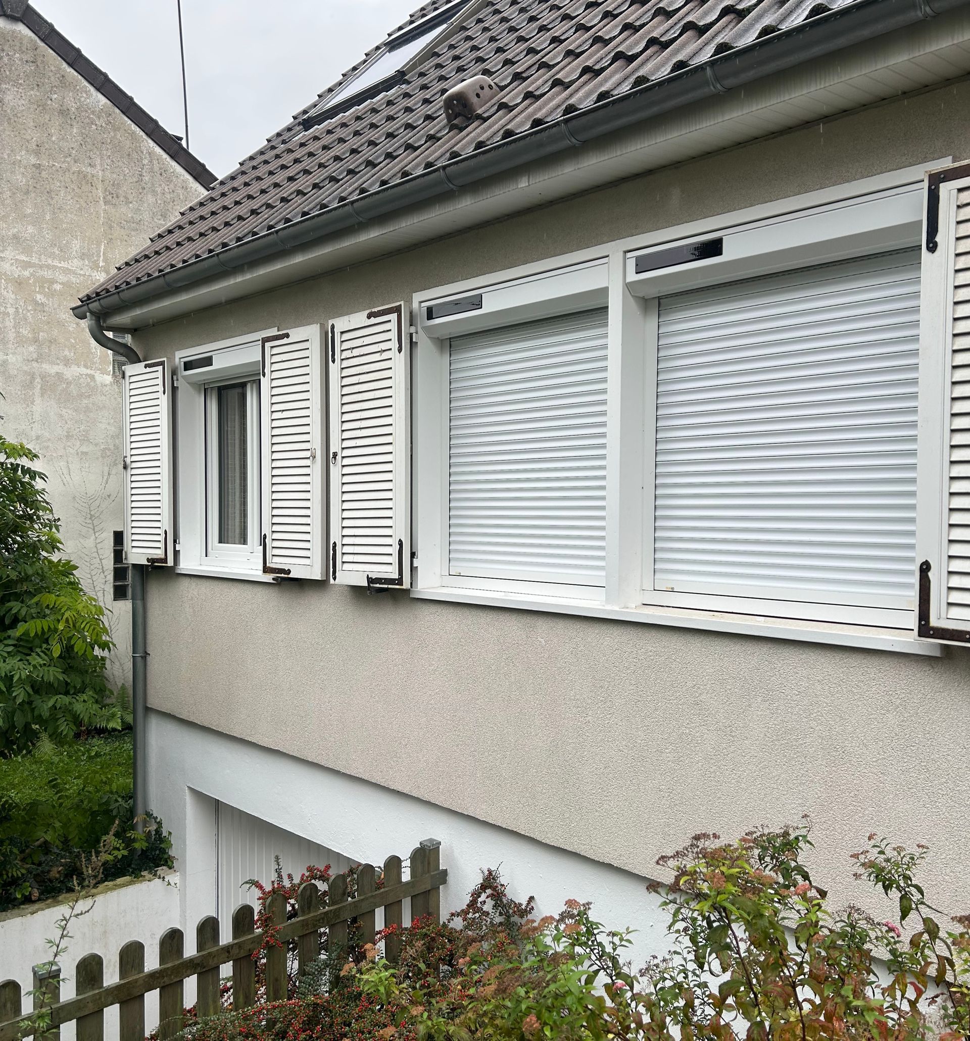 Bâtiment blanc avec volets, deux fenêtres avec stores fermés et une petite fenêtre avec volets ouverts.