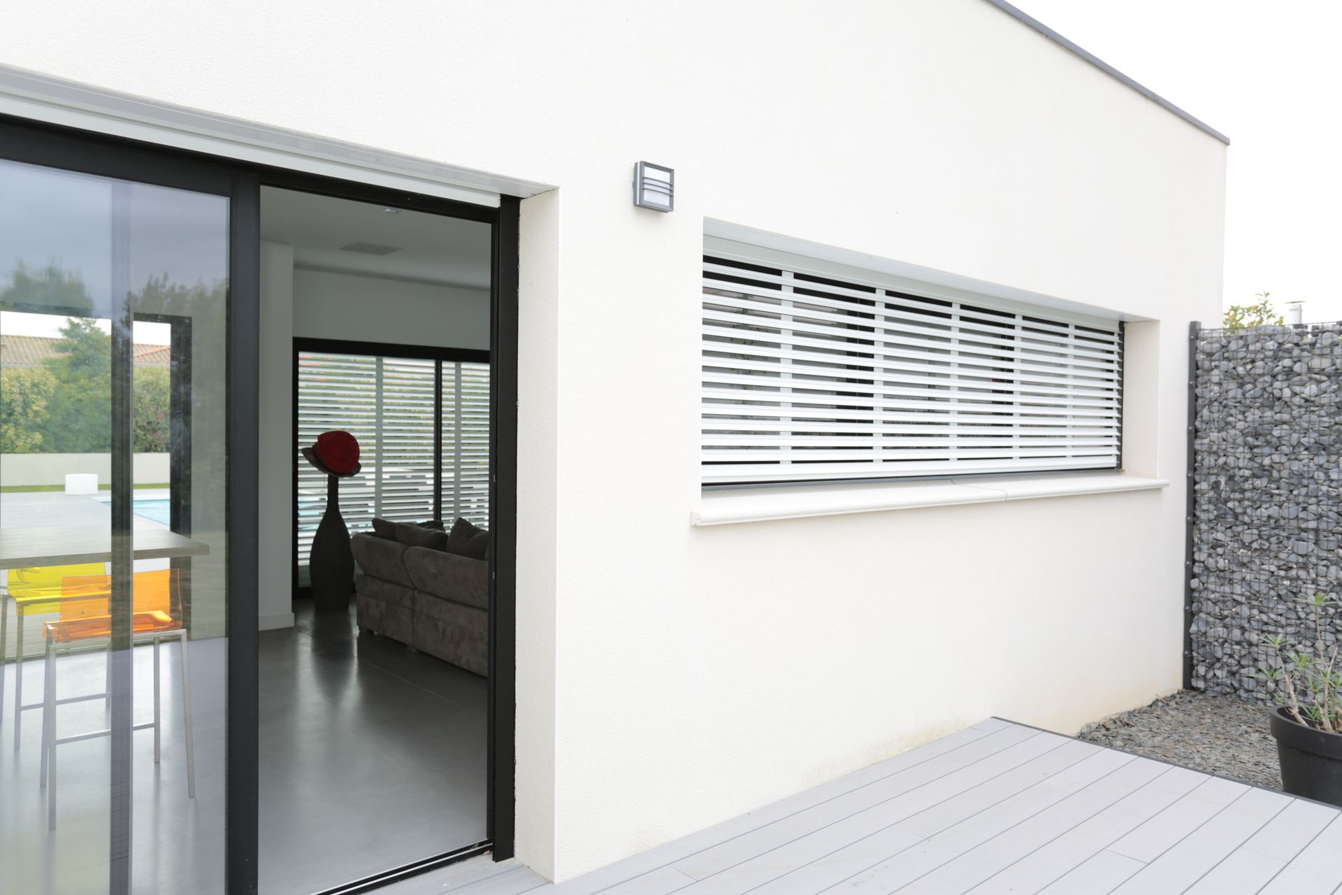 Extérieur d'une maison blanche moderne avec porte coulissante en verre et fenêtre à volets, à côté d'un mur de pierre et d'une terrasse.