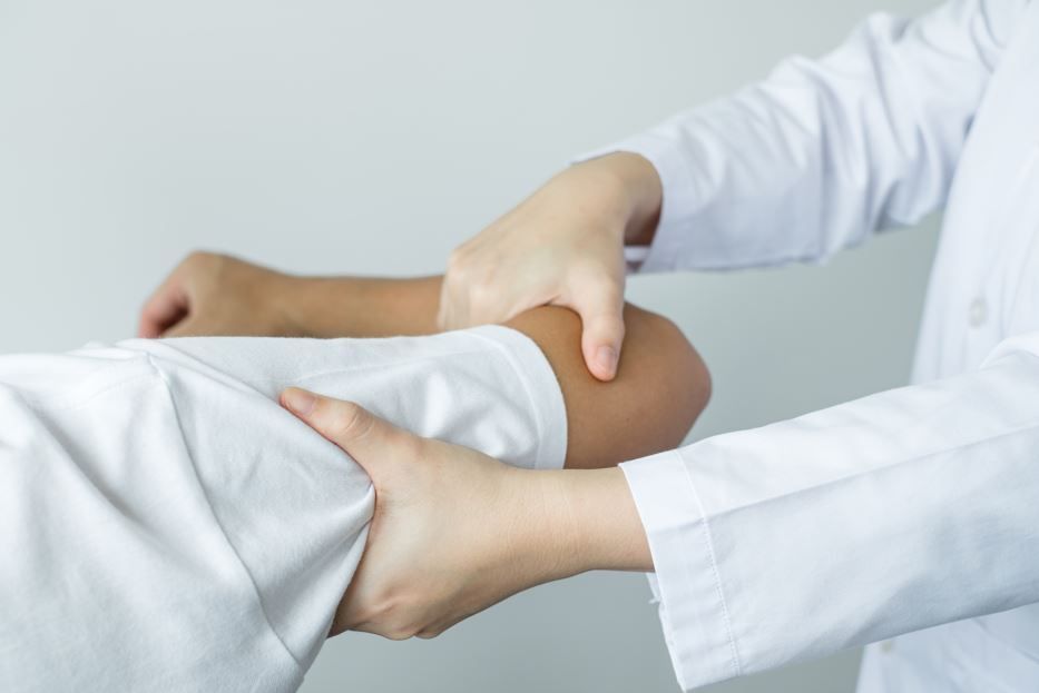 Médico manipulando el codo de un paciente, ambos vistiendo batas blancas, en un entorno clínico.
