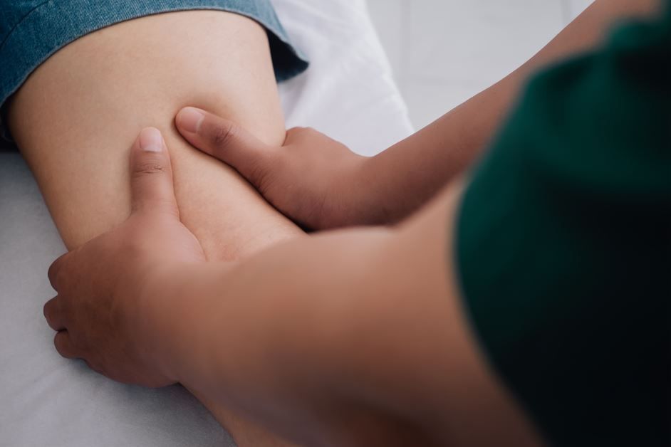 Manos masajeando la pierna de una persona. La persona lleva vaqueros azules, el entorno es blanco.