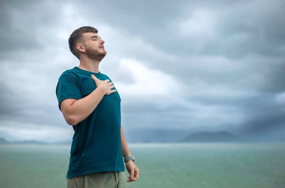 Hombre al aire libre con la mano en el pecho, mirando hacia arriba, cielo nublado y océano de fondo.
